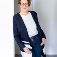 Frau in einem modernen Business-Outfit mit einer weißen Bluse und einem dunklen Blazer, die entspannt in einem hellen Raum steht und in die Kamera lächelt.
