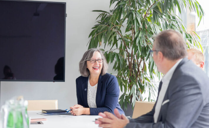 Foto einer Frau in einem Business-Meeting, die in einem Büro sitzt und mit Kollegen spricht, lächelt.