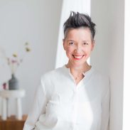 Foto einer Frau mit kurzen grauen Haaren und roten Lippen die in die Kamera lacht