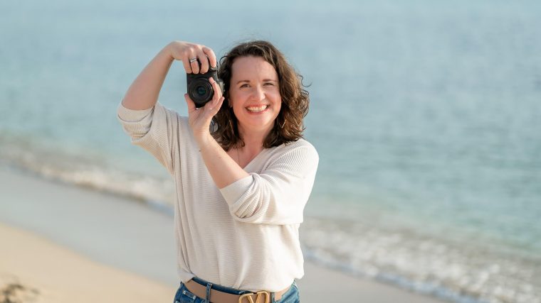 Professionelle Fotografin hält eine Kamera vor sich und lächelt, während sie am Strand steht und den Moment einfängt.