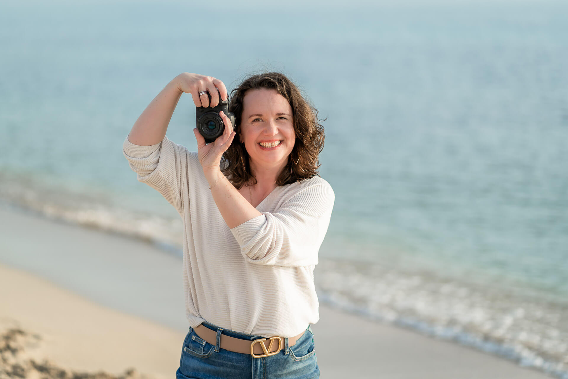 Professionelle Fotografin hält eine Kamera vor sich und lächelt, während sie am Strand steht und den Moment einfängt.