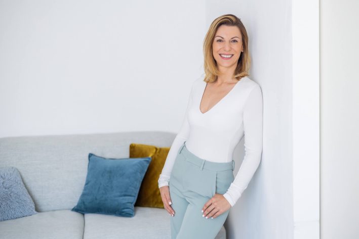 Foto einer Frau in einem weißen Oberteil und grauen Hosen, die an einer Wand lehnt und in die Kamera lächelt. Sie steht vor einer modernen Couch mit bunten Kissen.