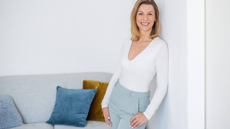 Foto einer Frau in einem weißen Oberteil und grauen Hosen, die an einer Wand lehnt und in die Kamera lächelt. Sie steht vor einer modernen Couch mit bunten Kissen.